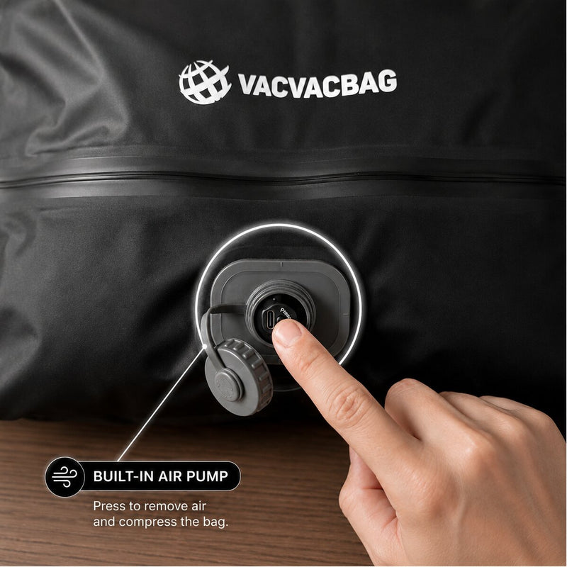 Close-up of a black vacuum bag with the VACVACBAG logo, showing a built-in air pump port that a finger is pressing to release air. A hand and part of a wooden surface are visible, with text reading 'BUILT-IN AIR PUMP' and 'Press to remove air and compress the bag.